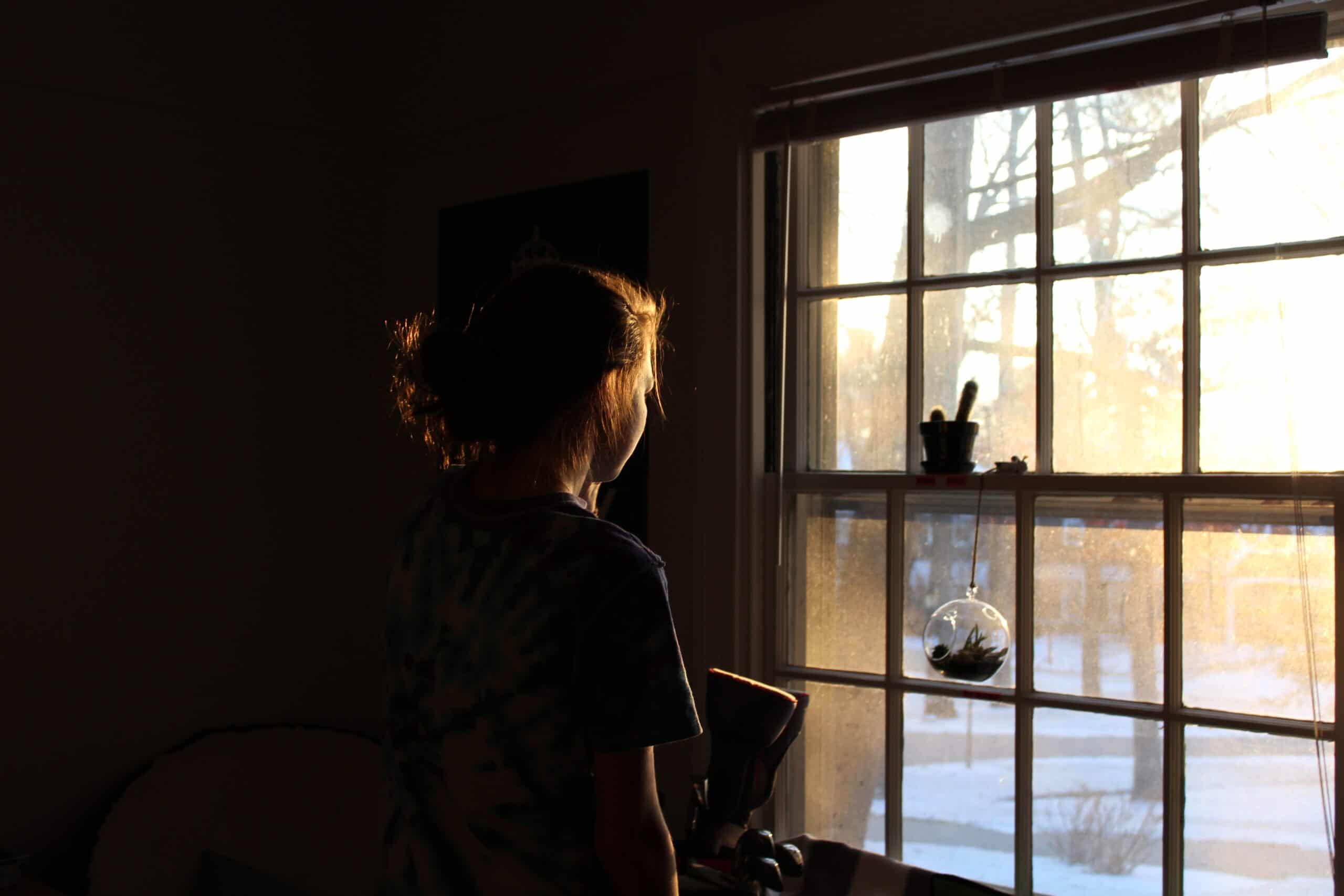 A person standing in a dark room looking out a window at the sunset. Trauma can feel isolating, but you're not alone. 